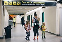 Das Bild zeigt eine Frau mit zwei kleinen Kindern an einem Flughafen. Alle tragen Rucksäcke.  