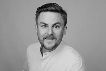 Porträt von Jasper Hoffmann. Ein Mann mit kurzen Haaren und Bart, der ein helles Oberteil trägt. Er lächelt leicht und blickt direkt in die Kamera vor einem neutralen grauen Hintergrund.