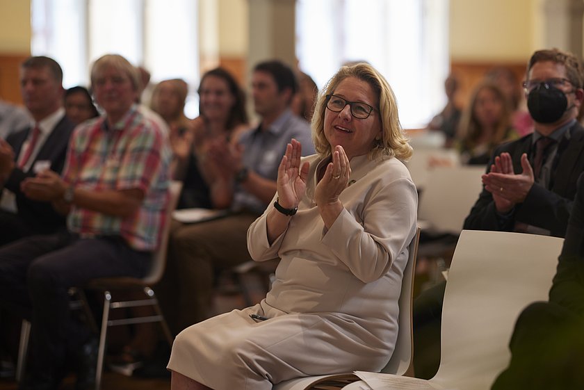 Eine Frau im Publikum applaudiert, während sie einem Vortrag in einem gut besuchten Veranstaltungsraum zuhört.