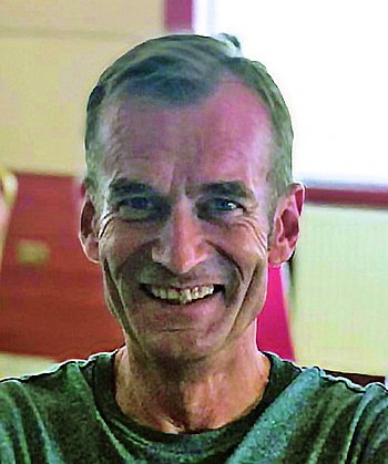 Ein lächelnder Mann in einem grünen Shirt, der vor einem roten Hintergrund sitzt.