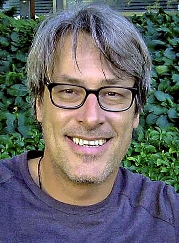 Porträt eines Mannes mit Brille, der draußen vor einer grünen Hecke lächelt.