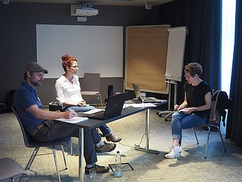 Drei Personen sitzen in einem abgedunkelten Raum an einem Tisch und nehmen einen Podcast auf. Zwei arbeiten an Laptops, während die dritte Person Notizen macht. Im Hintergrund sind eine Tafel und ein Flipchart erkennbar.