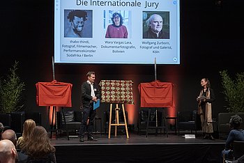 Ein Mann und eine Frau stehen auf einer Bühne. Im Hintergrund sieht man drei Staffeleien mit Bildern, die mit Tüchern verdeckt sind. Oben im Bild sieht man eine Präsentation, auf der Bilder von drei internationalen Jurymitgliedern zu sehen sind. 