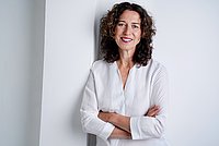 Portrait einer Frau mit schulterlangen braunen Locken und einem weißen Hemd. 