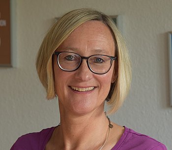 Eine Frau mit blonder Kurzhaarfrisur, die vor Schildern mit den Nachhaltigkeitszielen (SDGs) steht. Sie trägt eine violette Bluse und lächelt in die Kamera.