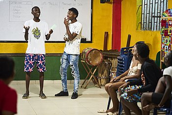 Zwei junge Männer stehen vor einer gelben Wand mit einer großen, weißen Tafel. An den Rändern des Bildes sitzen weitere Personen, die sich den beiden Männern zuwenden.