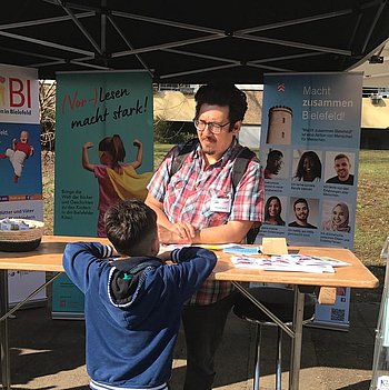 Ein dunkelhaariger Mann mit Bart und Brille im bunten Hemd steht an einem Stehtisch. Im Hintergrund befinden sich Rollups, auf denen "Vorlesen macht stark" und "Macht zusammen Bielefeld" steht. Ein Junge auf der anderen Seite des Tisches spricht mit dem Mann. 