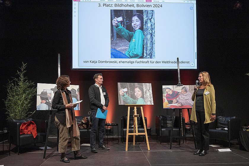 Drei Personen, zwei Frauen und ein Mann, befinden sich auf einer Bühne.  Im Hintergrund steht ein Foto auf einer Staffelei. Auf dem Foto ist ein junges Mädchen, das sich selbst fotografiert. 