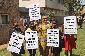 Eine kleinere Gruppe von Frauen und Männern posiert vor einem Gebäude und hält Schilder mit Forderungen zur Förderung der Frauenrechte und Bildung.