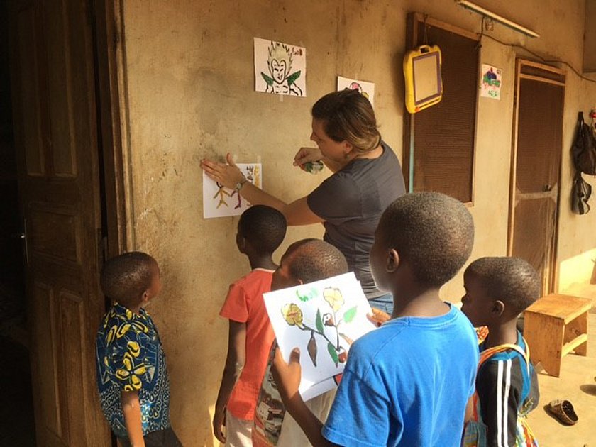 Eine Frau zeigt Kindern ein Poster an einer Wand. Die Kinder tragen bunte Kleidung und schauen aufmerksam zu.