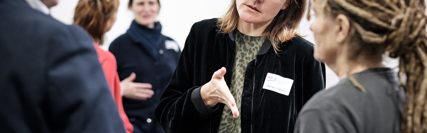 Eine Frau im schwarzen Blaser, einem Namenschild und mittellangen Haaren spricht mit einem Mann und einer Frau, die man nur von hinten bzw. von der Seite sieht. Der Mann trägt ein Jacket, die Frau hat einen grauen Pullover an und lange Dreadlocks. Die Frau mit dem Blazer gestikuliert mit der Hand. 