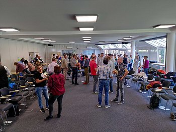 Ein heller Seminarraum mit großen Fenstern, in dem Menschen in kleinen Gruppen miteinander sprechen. Die Stühle sind zur Seite gestellt, und die Atmosphäre wirkt lebhaft und kommunikativ.