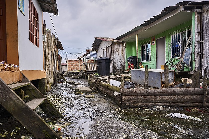 Das Bild zeigt einen durchnässten Weg an dessen Seiten kleine Hütten stehen. Auf der rechten Seite sitzt vor einer grünen Hütte eine Frau und ein Kind.