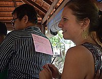 Eine lächelnde dunkelhaarige Frau klebt einen beschrifteten rosa Zettel auf den Rücken eines vor ihr stehenden Mannes. Der Mann trägt ein blau-weiß gestreiftes Hemd und hat dunkle Haare. 