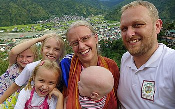 Eine Familie mit sechs Personen, bestehend aus zwei Erwachsenen, Mann und Frau und vier Kindern, lächelt in die Kamera. Im Hintergrund ist eine grüne Hügellandschaft mit vereinzelten Häusern zu sehen.
