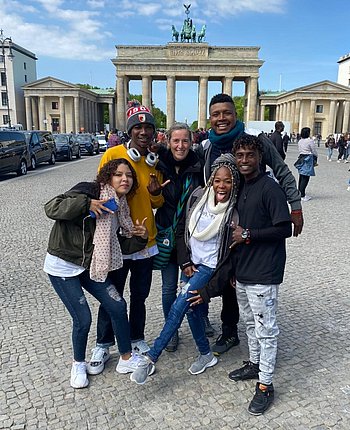 Fünf jugendliche Personen in sportlicher Kleidung und eine erwachsene Frau lächeln vor einem großen Tor in die Kamera.