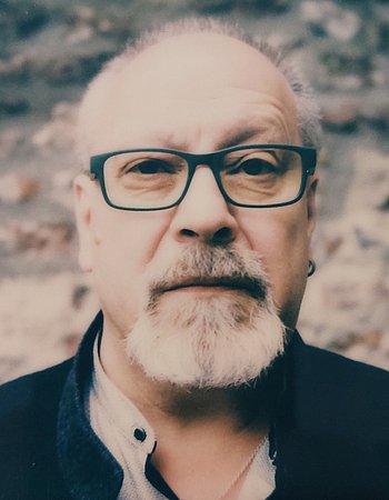 Das Bild zeigt einen Mann mit einem weiß-grauen Bart mit Brille. Er trägt einen dunkelblauen Pullover und darunter ein helles Hemd. 