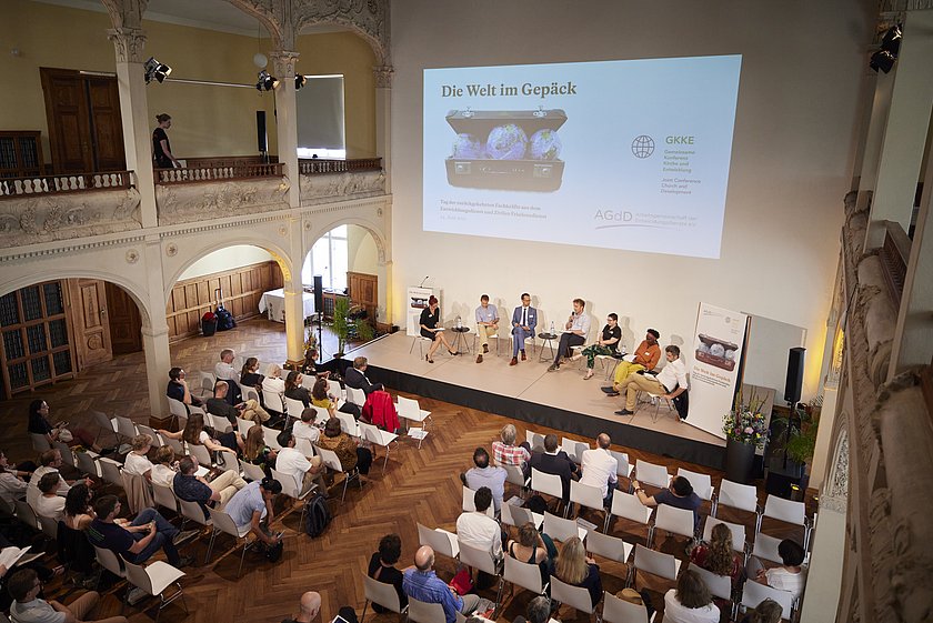 Eine breite Ansicht des Veranstaltungsraums mit einer großen Leinwand, auf der die Veranstaltung zu sehen ist.