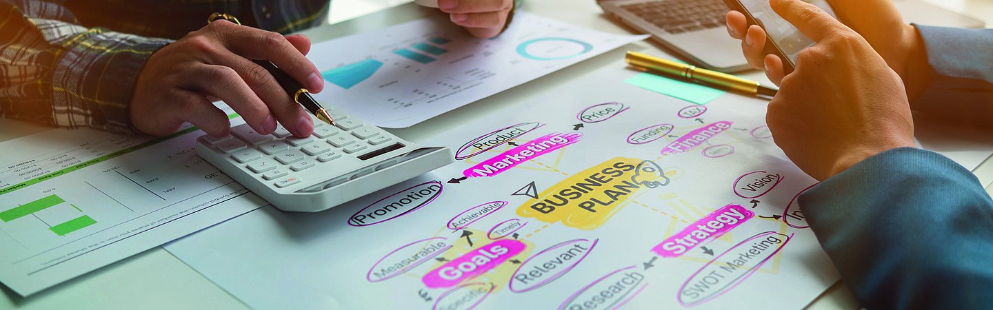 Zwei Personen sitzen an einem Tisch und beugen sich über ein großes Stück Papier. Auf dem Papier ist in der Mitte der Begriff "Business plan" und darum weitere Begriffe aus dem Marketing geschrieben. Von den Personen sind nur die Hände im Bild. Eine hält einen Becher in der Hand und tippt mit der anderen Hand auf einem Taschenrechner. Die zweite Person tippt auf einem Smartphone. Auf dem Tisch steht ein Laptop.