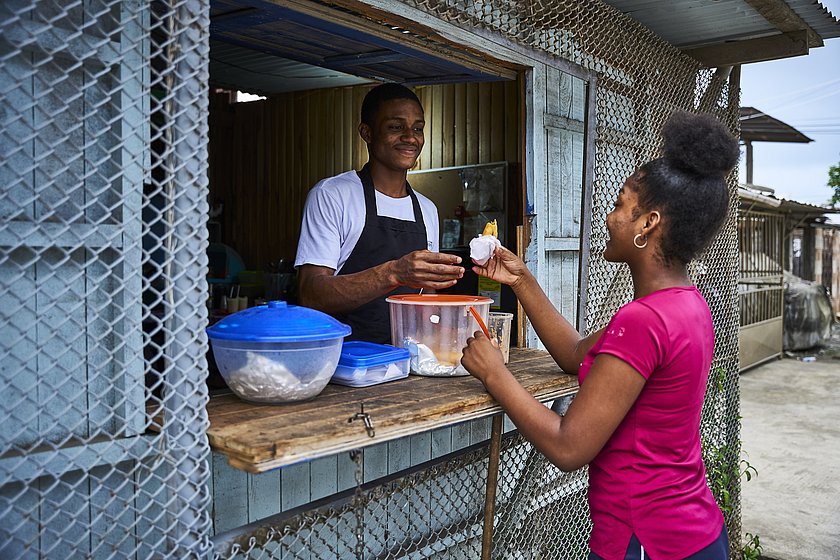 Ein Junge steht in einem Imbiss und überreicht einem Mädchen eine Teigtasche. 