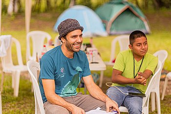 Ein lachender Mann mit Strohhut und grün-blauem T-Shirt und ein Junge mit grünem T-Shirt sitzen auf weißen Plastikstühlen und schauen in eine Richtung. Im Hintergrund sind Zelte zu sehen und weitere Plastikmöbel zu sehen..  