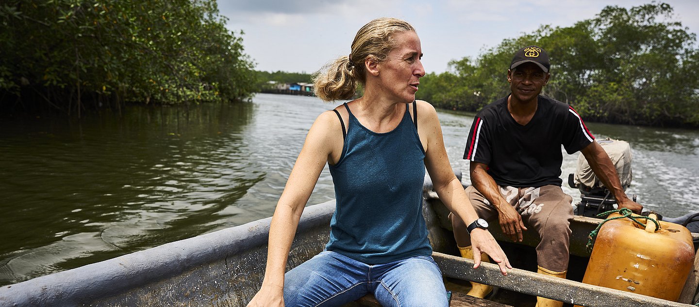 Eine Frau und ein Mann sitzen in einem Motorboot aus Holz auf einem Fluss. Entlang des Wassers sind Bäume.