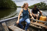 Eine Frau und ein Mann sitzen in einem Motorboot aus Holz auf einem Fluss. Entlang des Wassers sind Bäume.