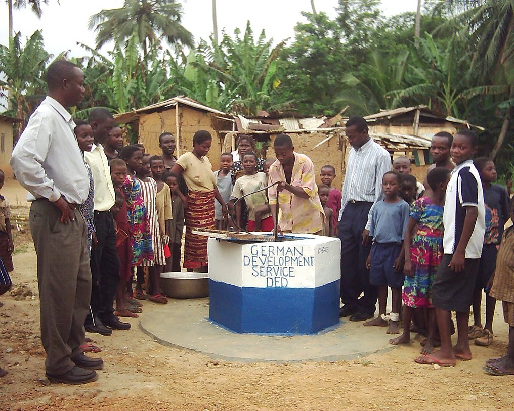 Die Bewohner des Dorfes stehen in einem Halbkreis um einen Brunnen herum.
