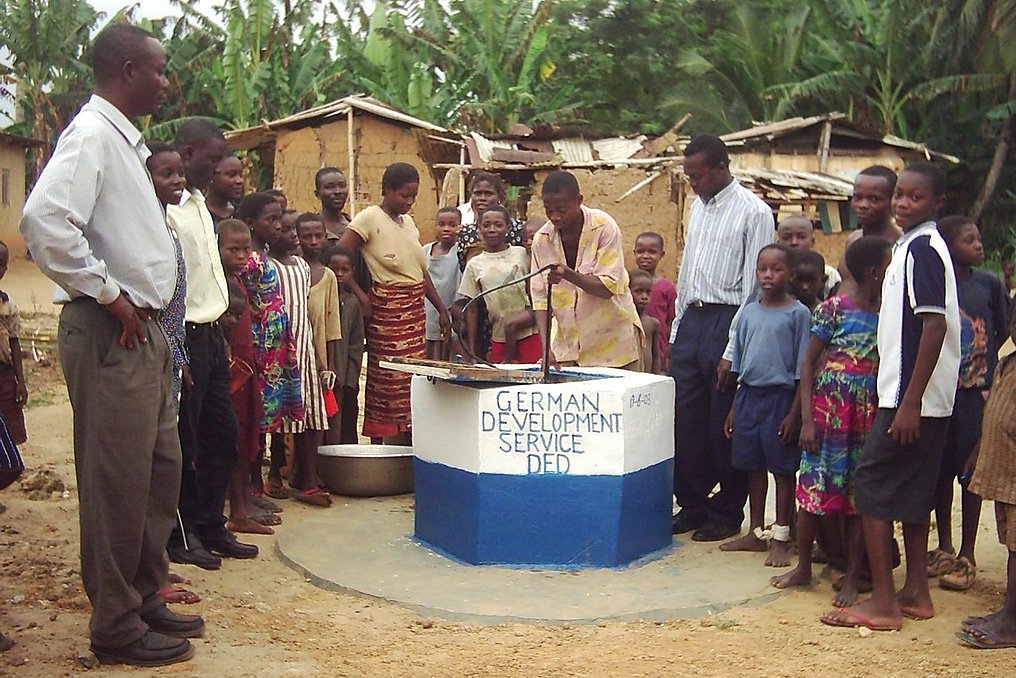 Die Bewohner des Dorfes stehen in einem Halbkreis um einen Brunnen herum.