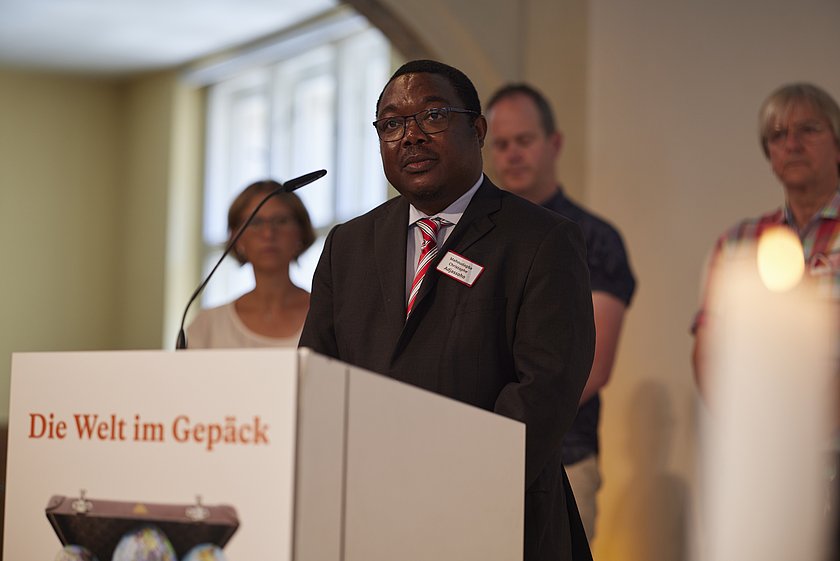 Ein Mann in einem dunklen Anzug spricht an einem Rednerpult mit der Aufschrift "Die Welt im Gepäck". Im Hintergrund stehen weitere Personen, die aufmerksam zuhören.