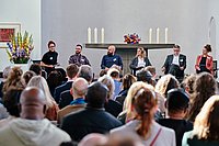 Ein Blick über die Köpfe eines Publikums hinweg auf eine Bühne, auf der mehrere Personen sitzen, umgeben von Kerzen und Dekoration.