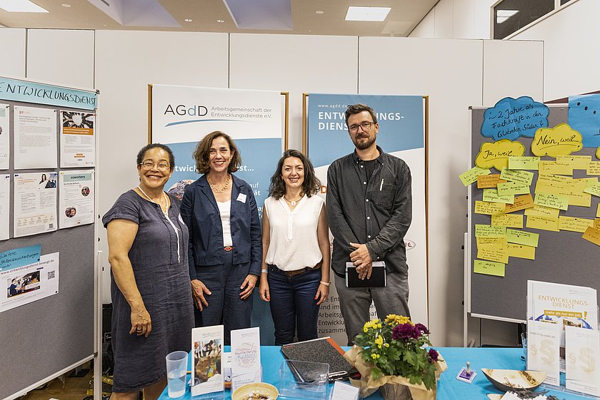 Vier Personen stehen gemeinsam an einem Messestand und posieren für ein Foto. Der Stand ist mit Infopostern, Bannern und einem Tisch mit Broschüren sowie einem Blumenarrangement dekoriert. Im Hintergrund sind weitere Messematerialien und eine Pinnwand mit Karten zu sehen.