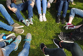 Kreis aus Jugendlichen, die auf einer Wiese stehen und verschiedene Schuhe tragen, im Fokus die Bodenperspektive.