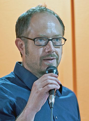 Ein Mann spricht in ein Mikrofon, während er nachdenklich zur Seite schaut. Im Hintergrund ist eine gelbliche Wand zu sehen.