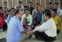 Eine Gruppe von Menschen sitzt im Kreis und beobachten einen Mann, den Entwicklungshelfer, der mittig im Bild sitzt. Er unterhält sich mit der Person rechts von ihm.