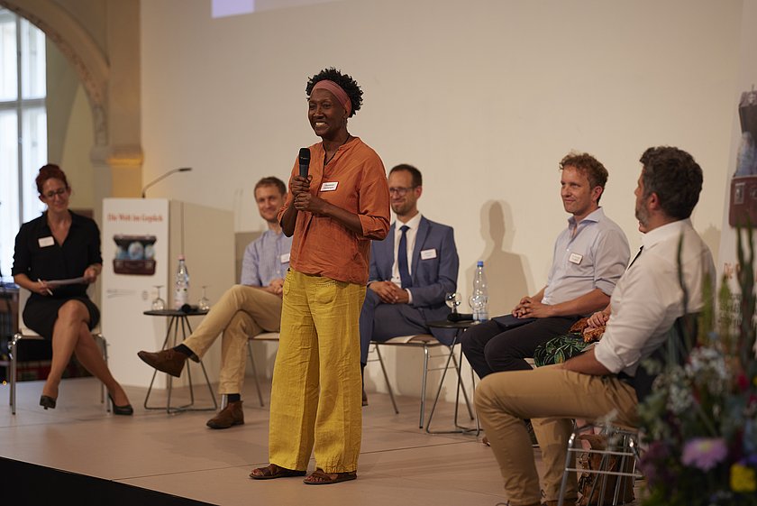ine stehende Person hält eine Rede, umgeben von einer Gesprächsrunde.