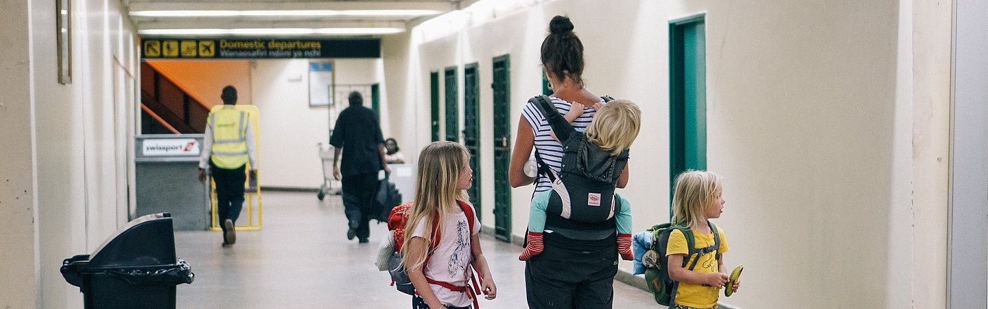 Das Bild zeigt eine Frau mit zwei kleinen Kindern an einem Flughafen. Alle tragen Rucksäcke.  