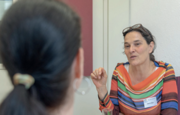 Eine Frau in einem bunten Oberteil spricht gestikulierend mit einer anderen Person, die von hinten zu sehen ist. Ein ein Beratungsgespräch in einem hellen Raum.