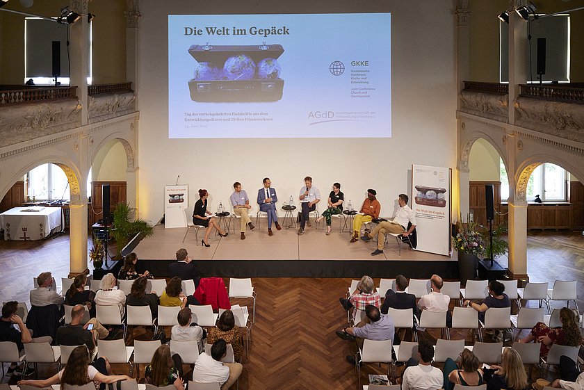 Eine breite Ansicht des Veranstaltungsraums mit einer großen Leinwand, auf der die Veranstaltung zu sehen ist.