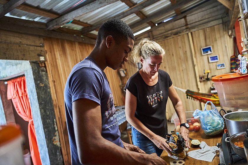 Ulrike Purrer steht mit einem jungen Manna an einem Holztisch. Auf dem Holztisch liegt Gemüse sowie eine Herdplatte. 