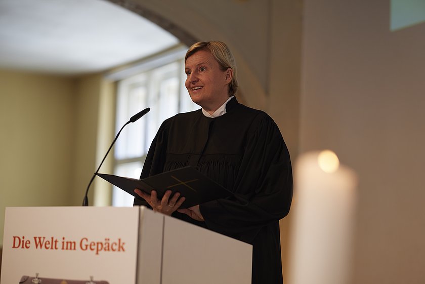 Eine Frau hält eine Rede an einem Pult mit einem Banner, das den Titel "Die Welt im Gepäck" zeigt.