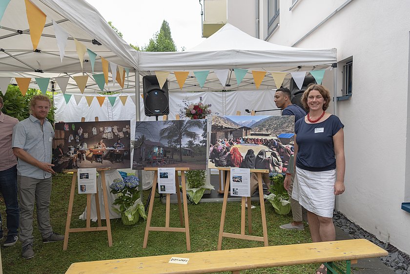 Eine Ausstellung im Freien unter einem Festzelt mit bunten Wimpeln. Drei großformatige Fotografien auf Staffeleien zeigen verschiedene Szenen aus unterschiedlichen Kulturen. Links steht ein Mann neben einem Bild, rechts eine Frau, beide lächeln. Im Hintergrund sind Blumenarrangements, ein Lautsprecher und weitere Besucher zu sehen.