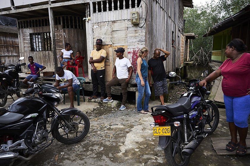 Ulrike Purrer steht rechts im Bild angelehnt an eine Holzhütte und spricht mit einem Mann, der sich den Kopf reibt. Um die Holzhütte versammelt sind verschiedene Menschen. Davor stehen Motorräder.
