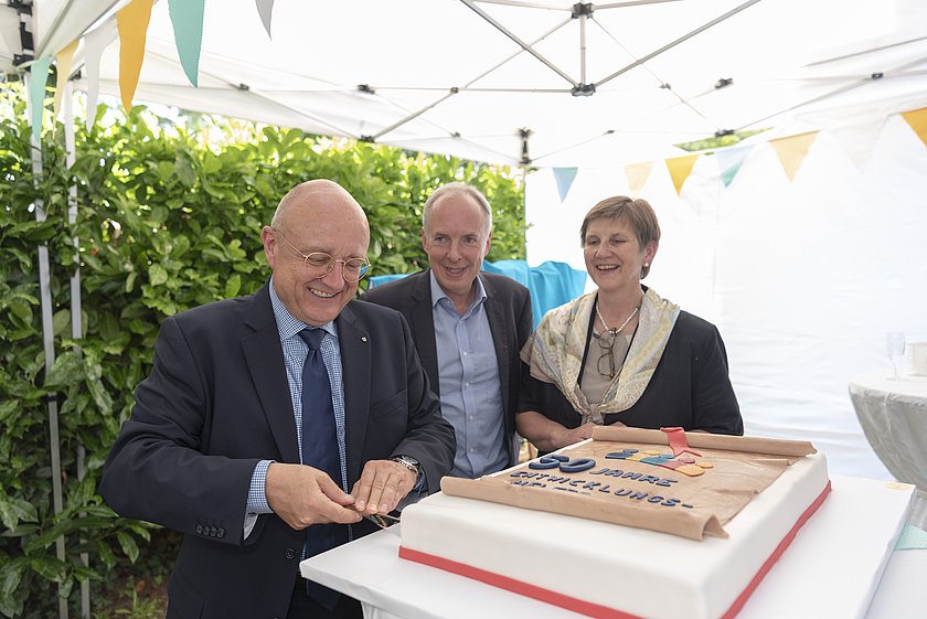 Zwei Männer und eine Frau in festlicher Kleidung stehen vor einer Torte. 
