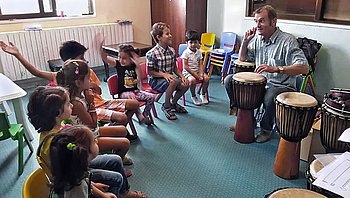 Kinder sitzen in einem Kreis und spielen Trommeln in einem hellen Raum.