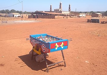 Ein bunter Tischkicker steht allein auf einer trockenen, sandigen Fläche. Im Hintergrund sind Gebäude und eine Moschee mit Minaretten zu erkennen.