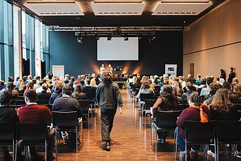 Ein Konferenzraum mit vielen Sitzreihen, in denen Teilnehmer einer Präsentation folgen. Ein großes Display ist im Hintergrund sichtbar.