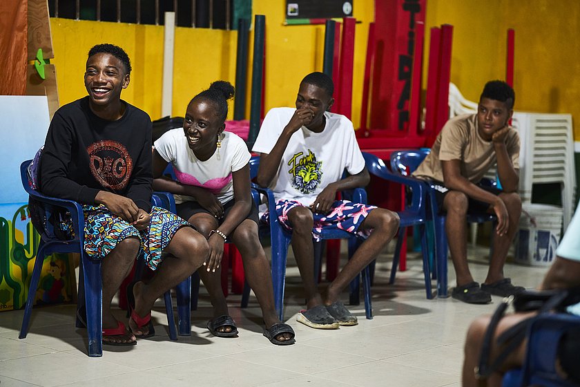 Drei junge Menschen sitzen mittig des Bildes auf Plastikstühlen. Alle drei lachen und beugen sich dabei leicht nach vorne.