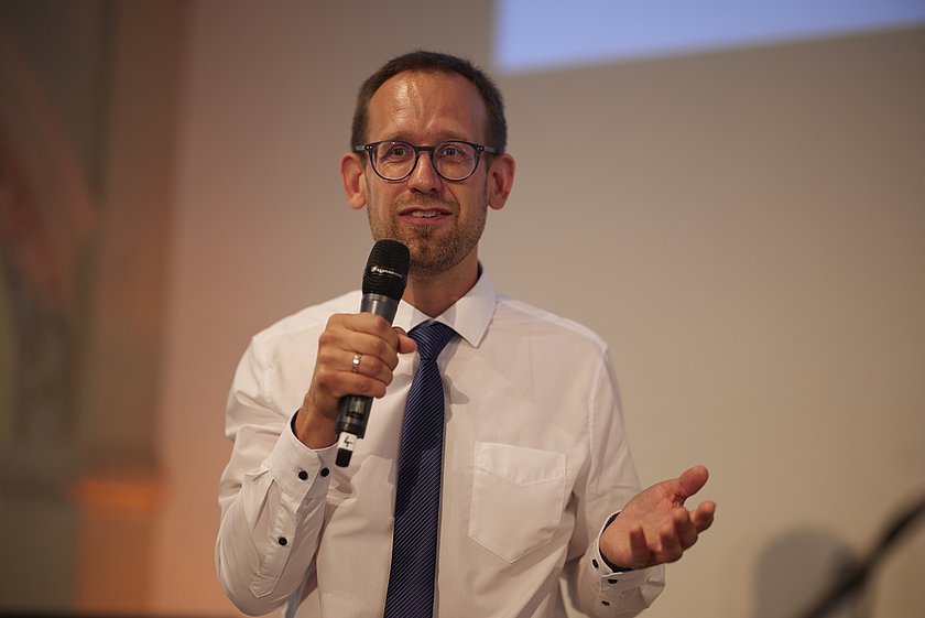 Ein Mann spricht mit einem Mikrofon vor einem Publikum.