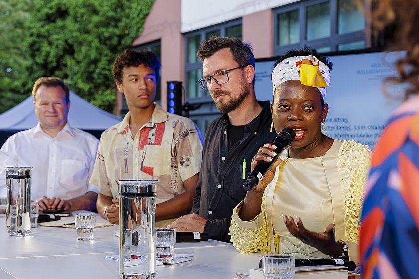 Vier Personen sitzen an einem Podiumstisch während einer Diskussion im Freien. Eine Frau in gelber Kleidung mit einem Mikrofon spricht, während die anderen aufmerksam zuhören. Im Hintergrund sind ein Gebäude und grüne Bäume sichtbar.